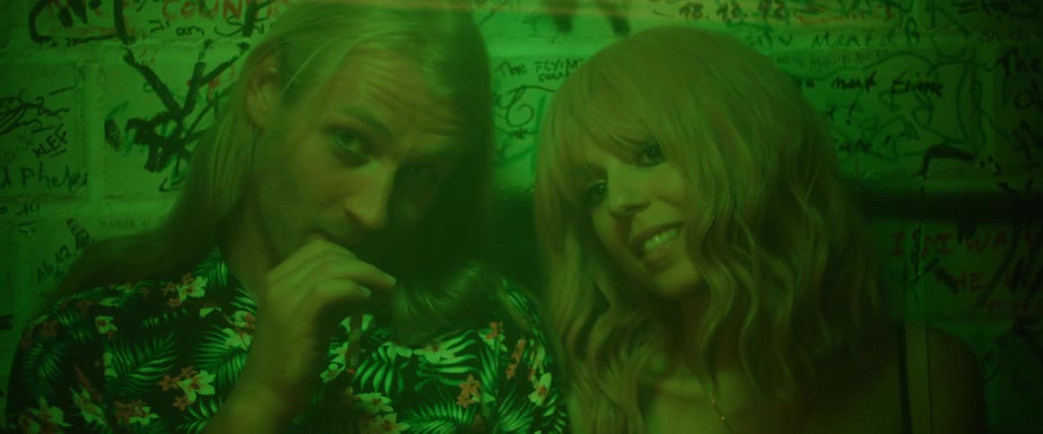A man and woman sit against a graffiti covered wall and talk as the man offers his cigarette