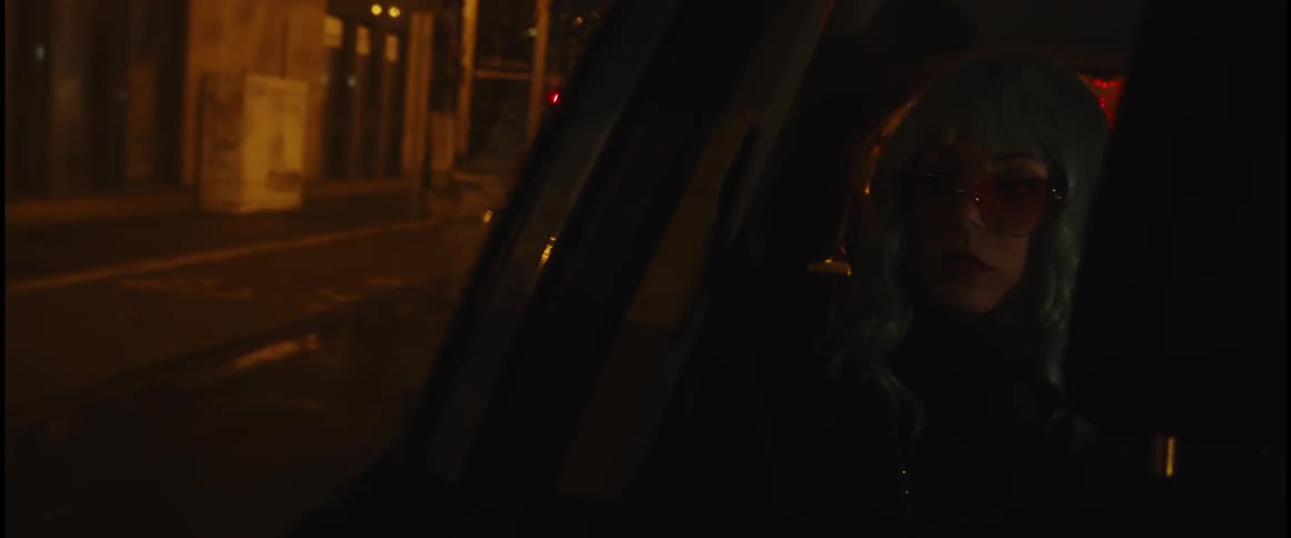 A woman with light green hair and big glasses is riding in an automobile and looking out the window at buildings and cars on the side of the road