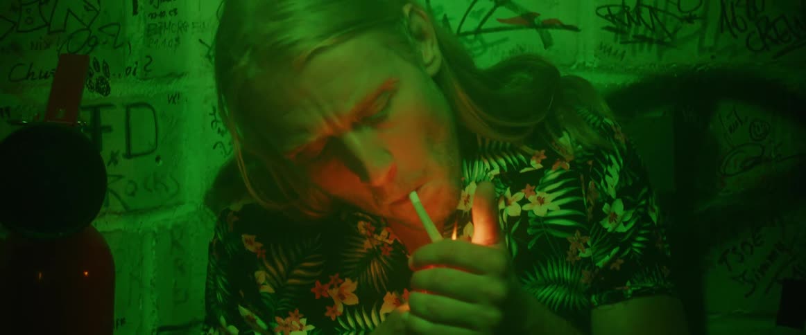 A man leans against a wall covered in graffiti and lights a cigarette and smokes