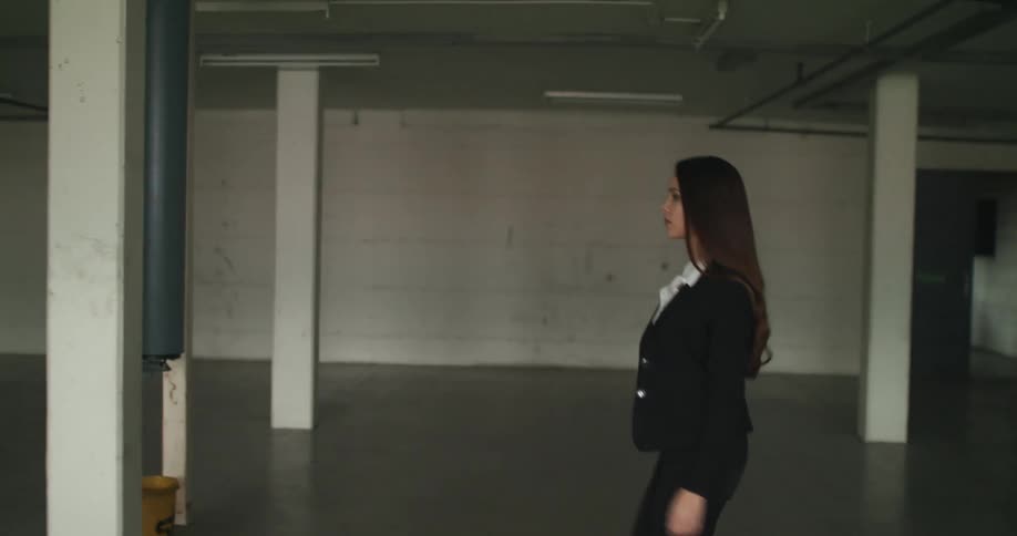 A woman with long dark hair and wearing a black pant suit is walking across an empty room