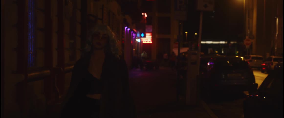 A woman wearing a wig slowly walks along a city street at nighttime