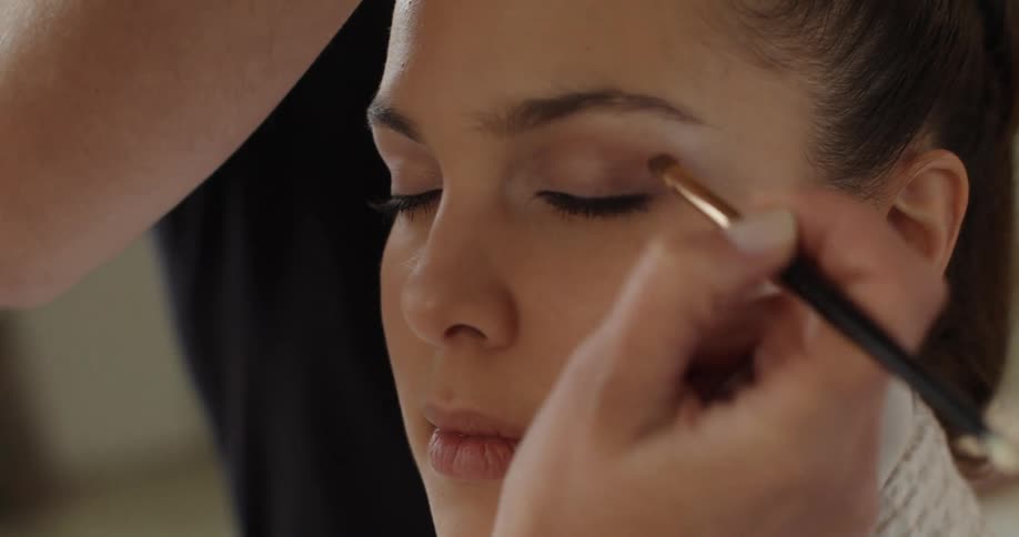 A woman is sitting as someone applies makeup and another person does her hair