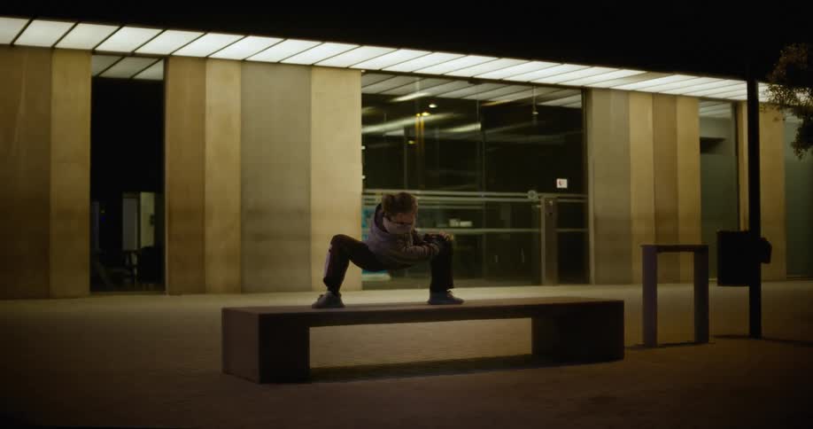 A woman in a wide squat position stretches on a bench outside at night