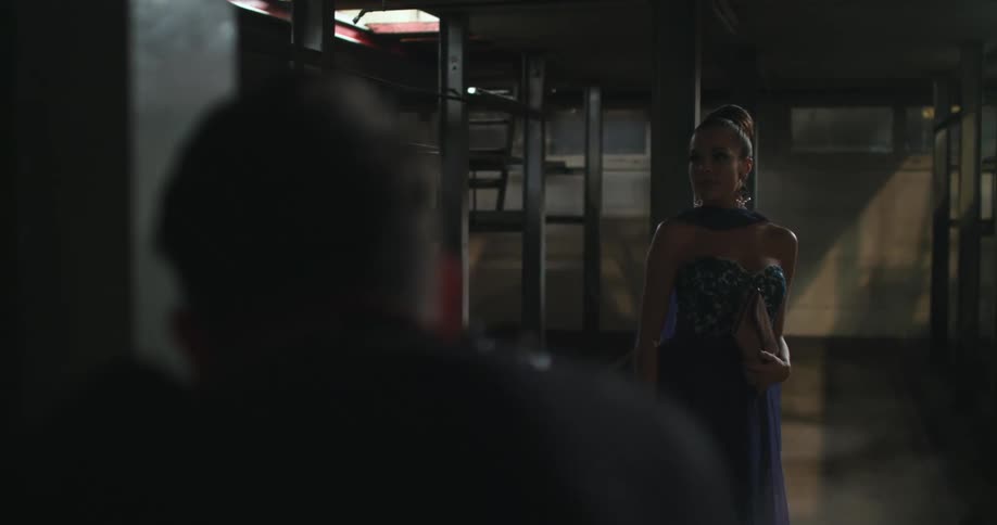 A woman wearing an evening dress and holding a purse is standing in an empty room as a man takes her picture