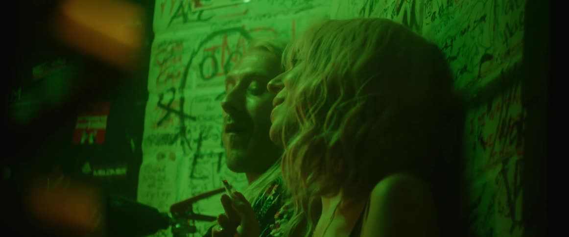 A man and woman lean against a wall covered in graffiti and smoke and the intoxicated woman leans on the man as she stumbles
