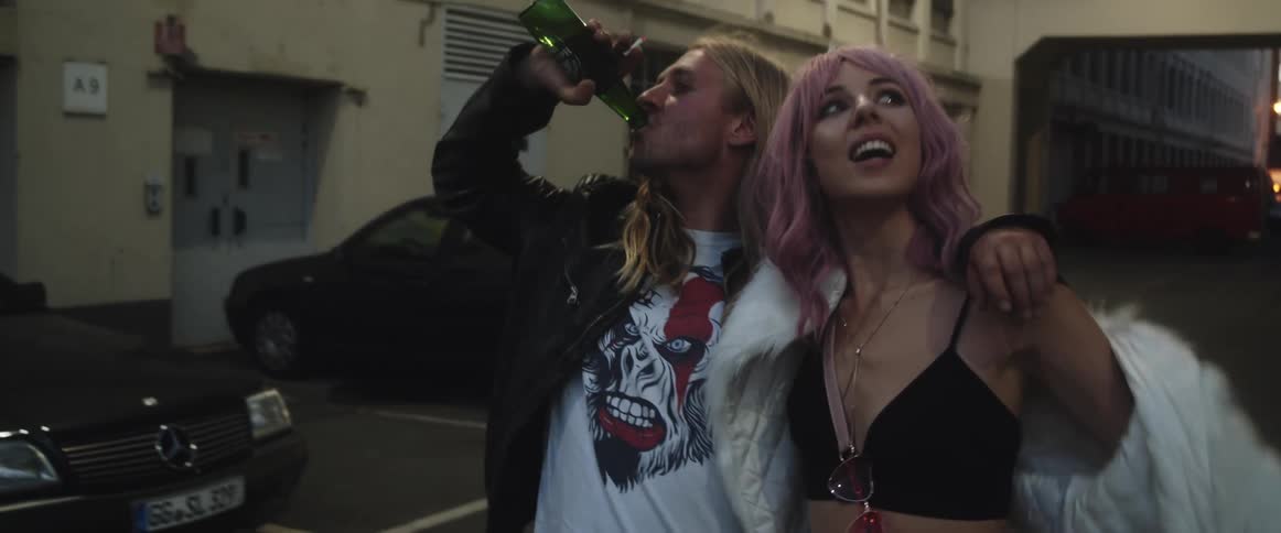 A man drinks wine from a bottle while walking with his girlfriend through a parking lot