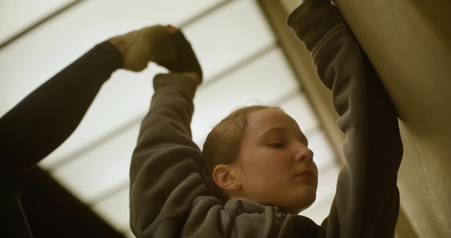 A teenage girl is practicing ballet as she leans her arm on the wall and holds her foot in the air above her head