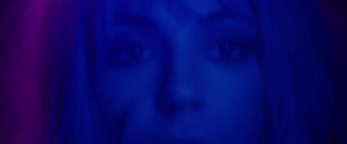 A purple light is shining on a blond woman who is looking up and takes off her glasses