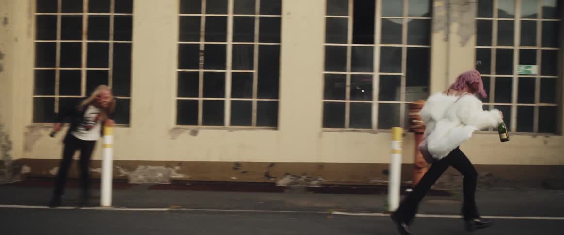 A pink haired woman wearing a white fur coat and a long haired man are running on a street each carrying a bottle and playing chase as he leaps over a post