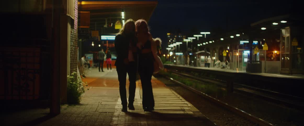 A couple stumbles as they walk down a city sidewalk drinking from a bottle