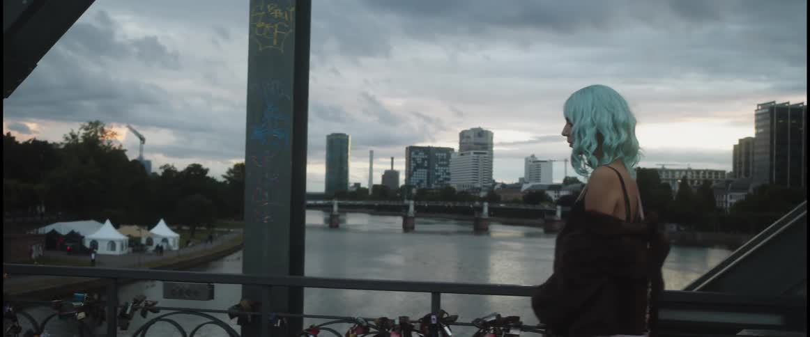 Young woman with bright blue hair walking on a bridge over a river in the city at evening