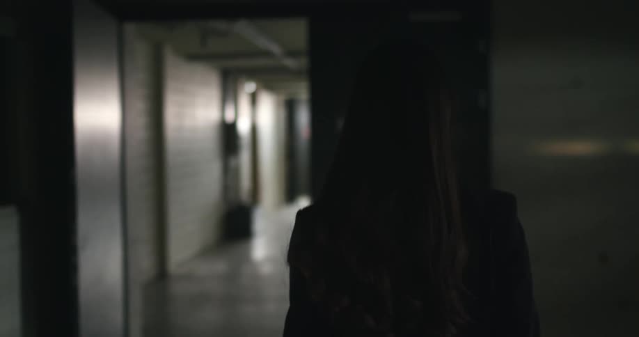 A woman with long dark hair walks down a corridor to a room with bright light shining in the windows