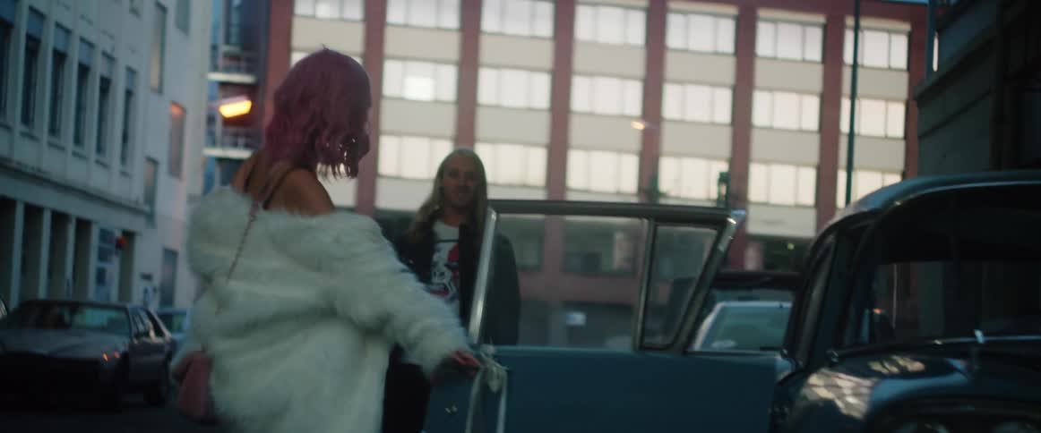 A pink haired woman wearing a white fur coat over a bikini top is carrying a wine bottle and walking on a street near an old car and she opens the door and gets inside as a man with her looks inside