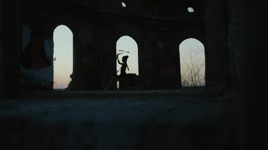 Silhouette of a ballerina sitting and waving a long ribbon in the air in a structure with arched windows and she stands to practice her dance