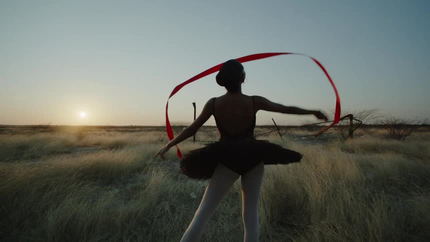 A ballerina is standing outside in a field doing a ribbon dance as the sun sets on the horizon
