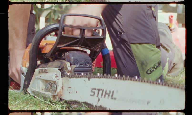 A kneeling man pulls the cord of a chainsaw resting on the ground to start it