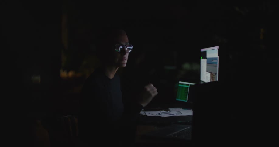 A woman is sitting at a desk with computer monitors and she looks at a laptop computer as she writes on paper