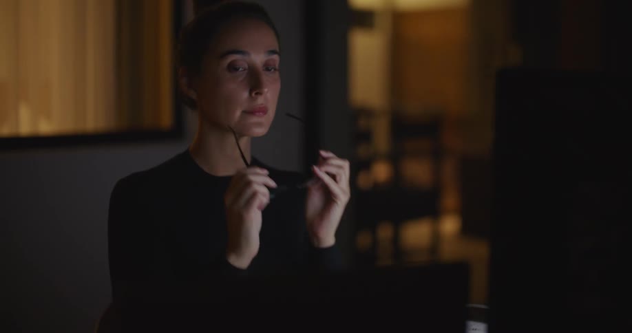 Woman working on laptop in dark room takes glasses off briefly to rub her eyes