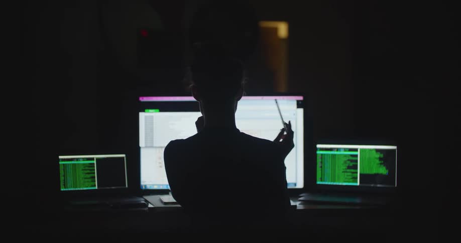 Woman is silhouetted against computer screens while working in dark room