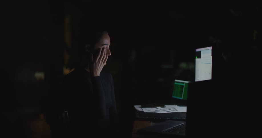 A woman removes her eyeglasses and rubs her eyes while looking at the screen of laptop on a desk in a dark office at night