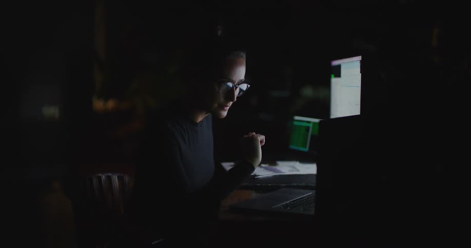 Woman works at desk in dark room consulting laptop screen and writing