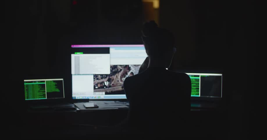Woman sits at a desk in a dark room and watches the graphics on three monitors