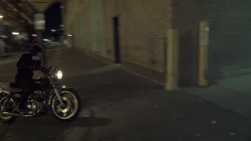 Man riding a motorcycle down a street and then turning  in between two buildings