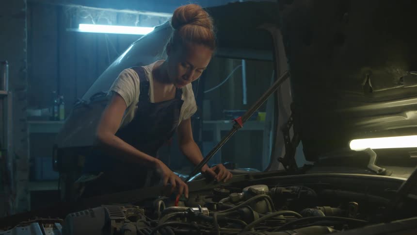 Woman examines car engine in a garage lit by a fluorescent bulb
