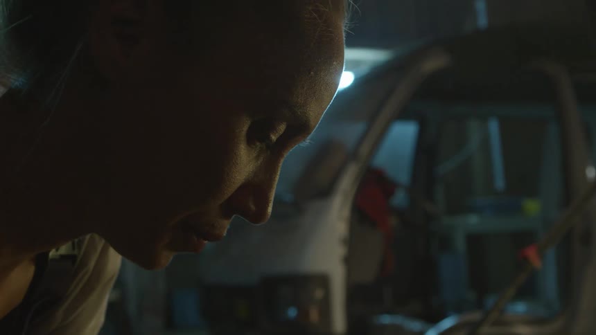 A woman is inside a garage bending down near an automobile and there are cans on a shelf