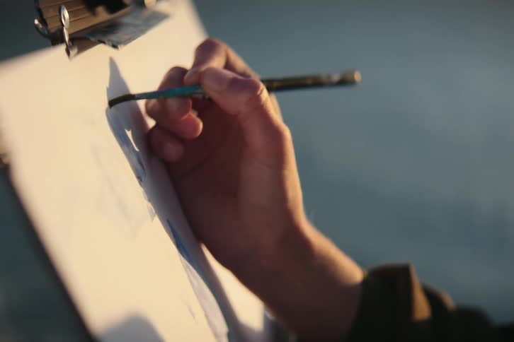 A hand holding a brush paints on paper canvas outside in the sunlight