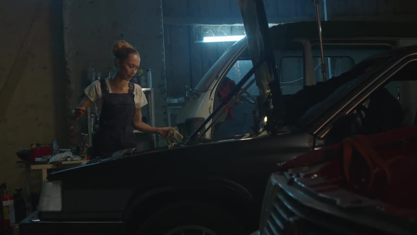 A female mechanic in a garage removes and examines the dipstick from the engine of a car before replacing it
