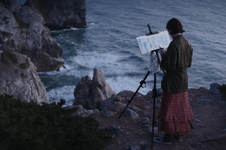 An artist is standing along the coast overlooking waves rolling in and splashing against the rocks below as she paints a picture on her easel