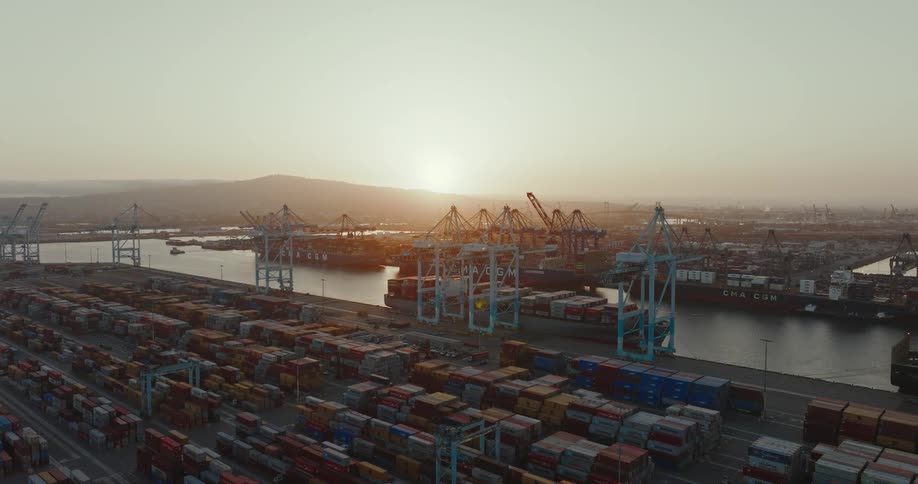 Cargo ships are at the loading dock and there are many shipping containers stacked along the dock as the sun shines over mountains in the distance