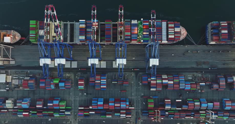 Cargo ships are at the loading dock as cranes move shipping containers onboard and trucks are carrying their loads