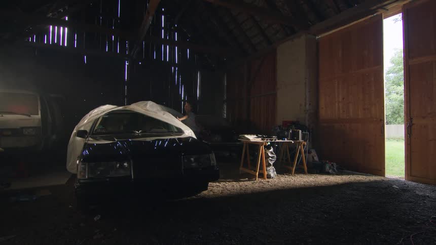 A woman is pulling the cover off of an automobile being stored in a barn as sunlight shines in from the door