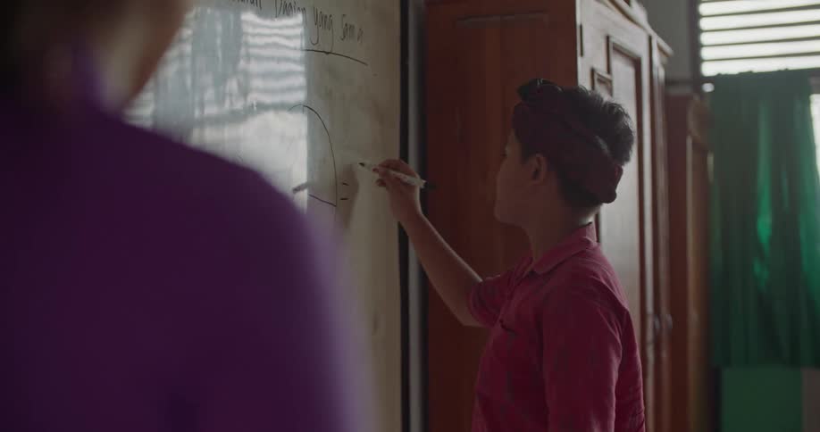 A student uses a marker to write on a whiteboard while a teacher observes