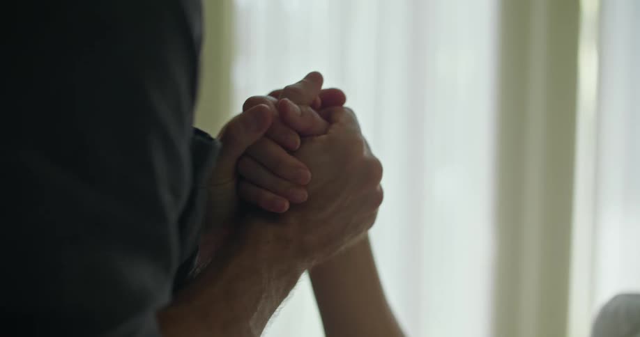 A woman is lying in bed and a man is holding her hand with both of his hands as he kisses her hand and smiles at her