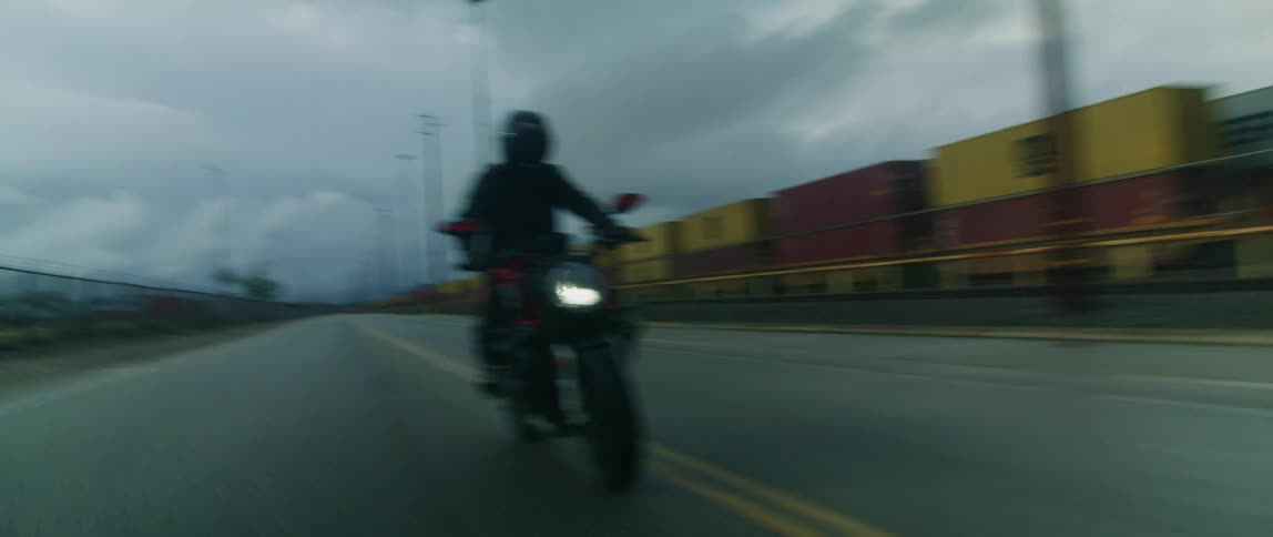 A cyclist dressed in black and wearing a black helmet is riding a motorcycle on a road alongside a train carrying cargo containers