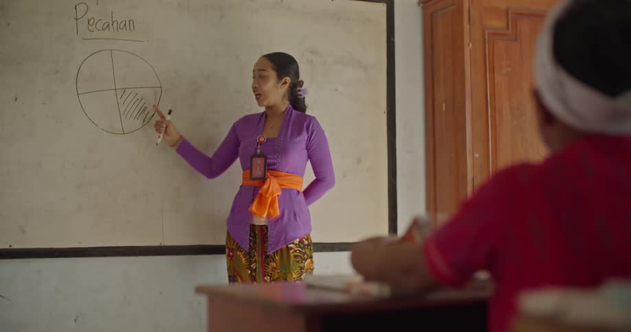 A teacher stands at the front of a classroom and points at a drawing on the board as a child sits at his desk flipping a book