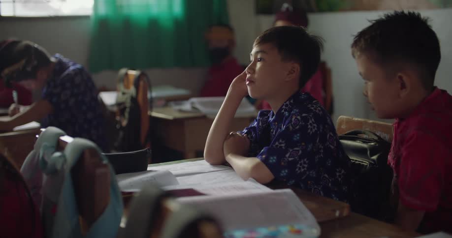 Student sitting at desk talks as others in classroom work and listen at their desks