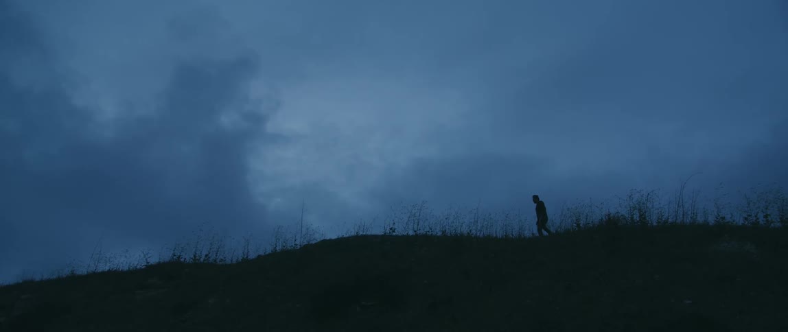 The silhouette of a person walking across the top of a grassy hill in on a cloudy evening