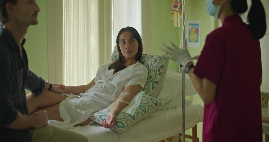 A woman is hooked up to an iv as a man sits on the bed holding her hand and a doctor speaks to them