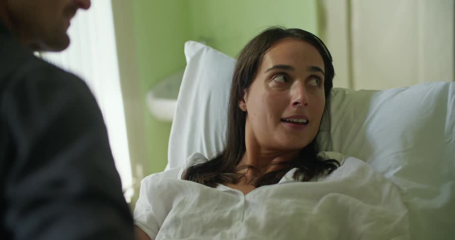 A woman is sitting up in bed with a pillow behind her head as she speaks and her husband holds her hand