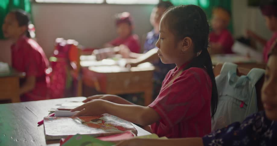 Young children sitting at desks in a classroom flip open books and look to the front of the room