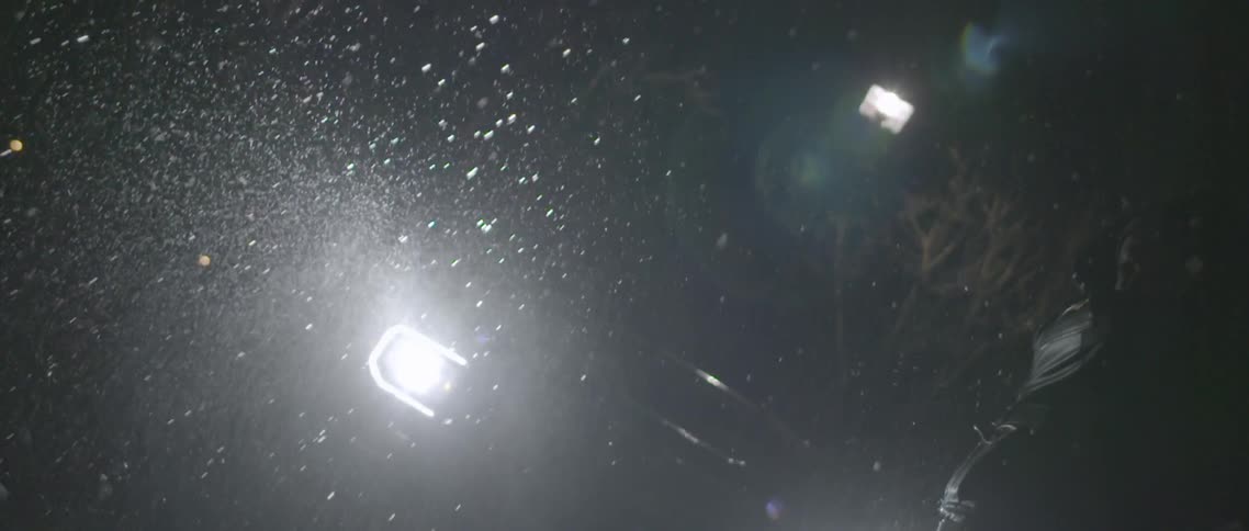 At night under bright lights a man swings a bat and shatters a water bottle