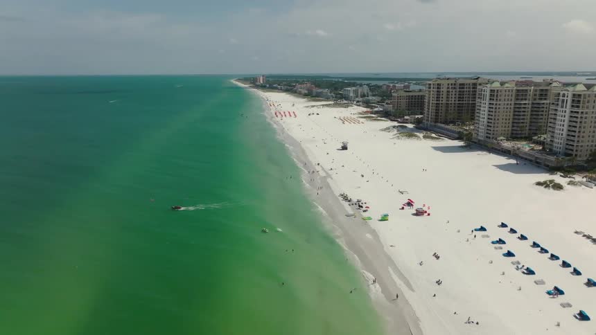 Sprawling hotel buildings line a sunny Florida beach as tourists watch the boats on the water