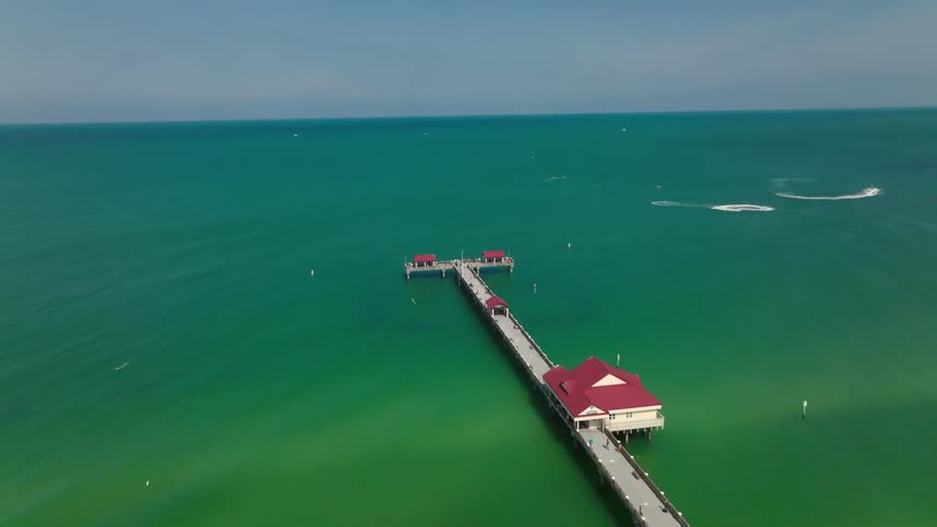 Drone shot following a long pier on a sunny Florida beach, flanked by tourists and boats