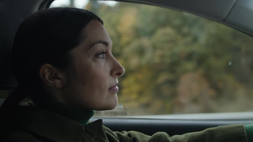 A woman is driving a car and trees can be seen from her drivers side window