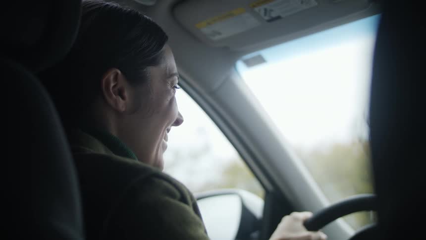 A woman is driving a car smiling and trees can be seen from the window