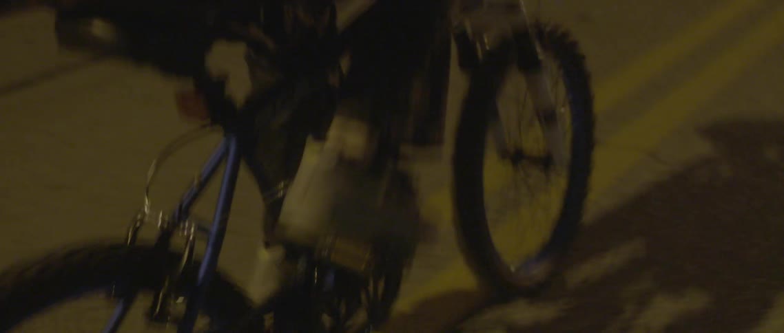 A boy is racing his bike down a street at night looking back frequently